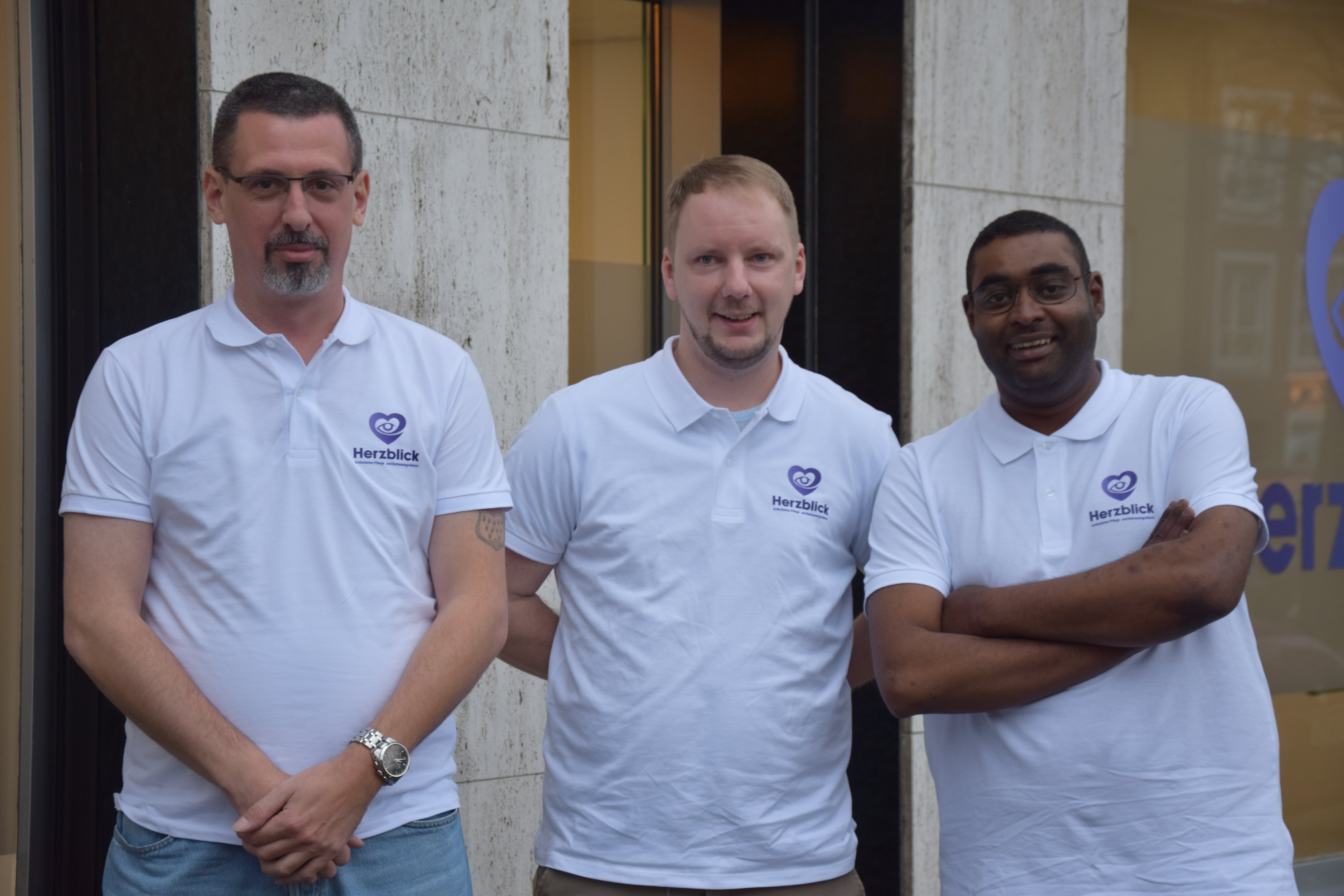 Drei Mitarbeitende des Pflegedienstes Herzblick stehen in weißen Poloshirts vor dem Eingangsbereich des Gebäudes.